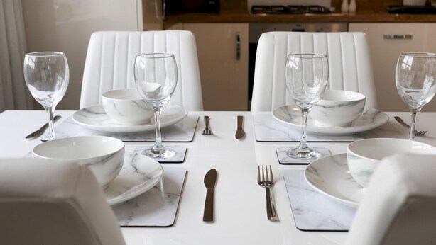 Une table à manger dressée avec de l’assiette blanche, des bols et des verres à vin, entourée de chaises blanches.