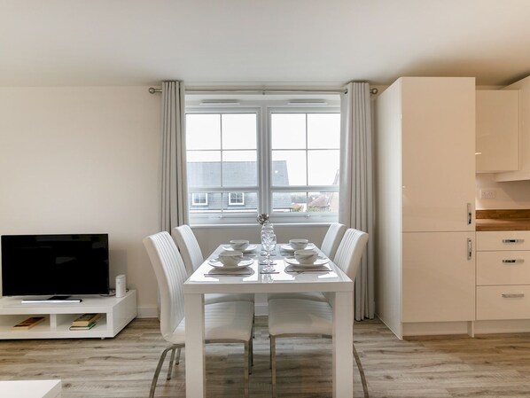 A modern dining area with a white table set for four, a flat-screen TV, and a kitchen with white cabinets.