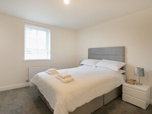 A bedroom with a bed, a bedside table with a lamp, a radiator, and a window with blinds.