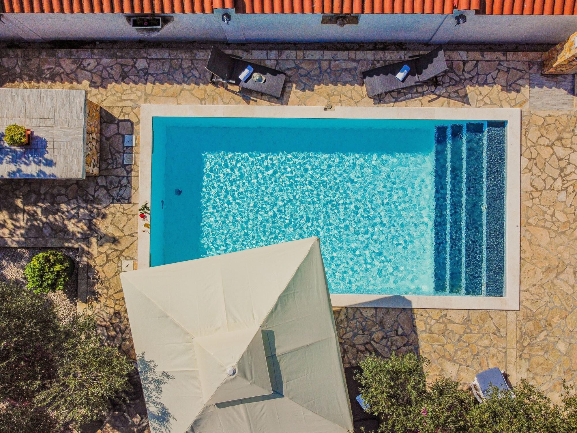Une vue aérienne d’une piscine rectangulaire à l’eau d’un bleu limpide, entourée de murs en pierre et surmontée d’un toit en tuiles.