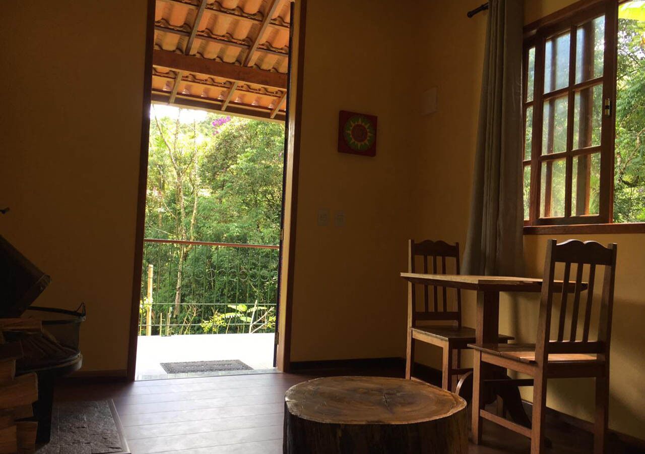A room with a wooden table and chairs, a small round table, a window with curtains, and a view of greenery outside.