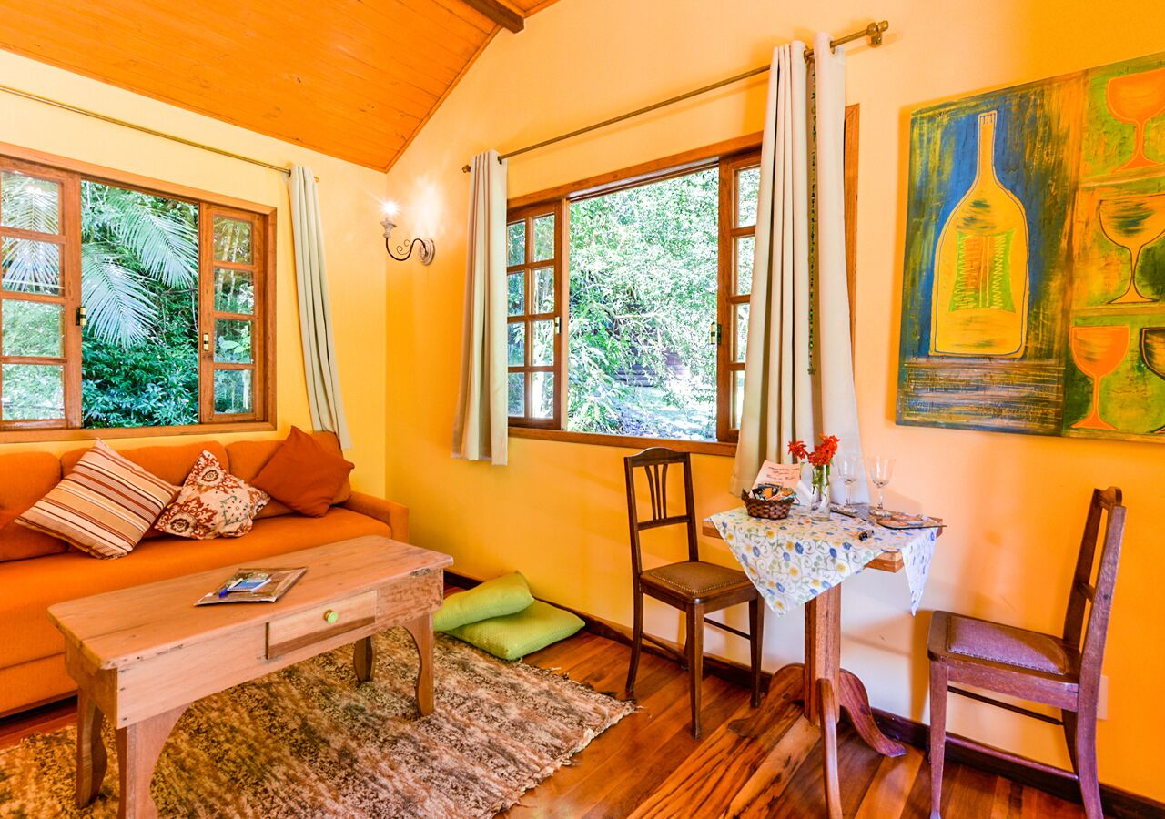 A cozy living room with a sofa, chairs, a coffee table, and a dining table set for two. Large windows provide natural light and a view of greenery outside.