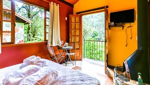 A bedroom with a bed, a small table, a chair, a TV, and a balcony with a view of greenery. - Amadamata Refúgio Ecológico (Nova Friburgo)