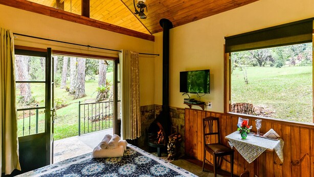 A cozy room with a fireplace, a bed with a patterned bedspread, a dining table with a vase, and a television. - Amadamata Refúgio Ecológico (Nova Friburgo)