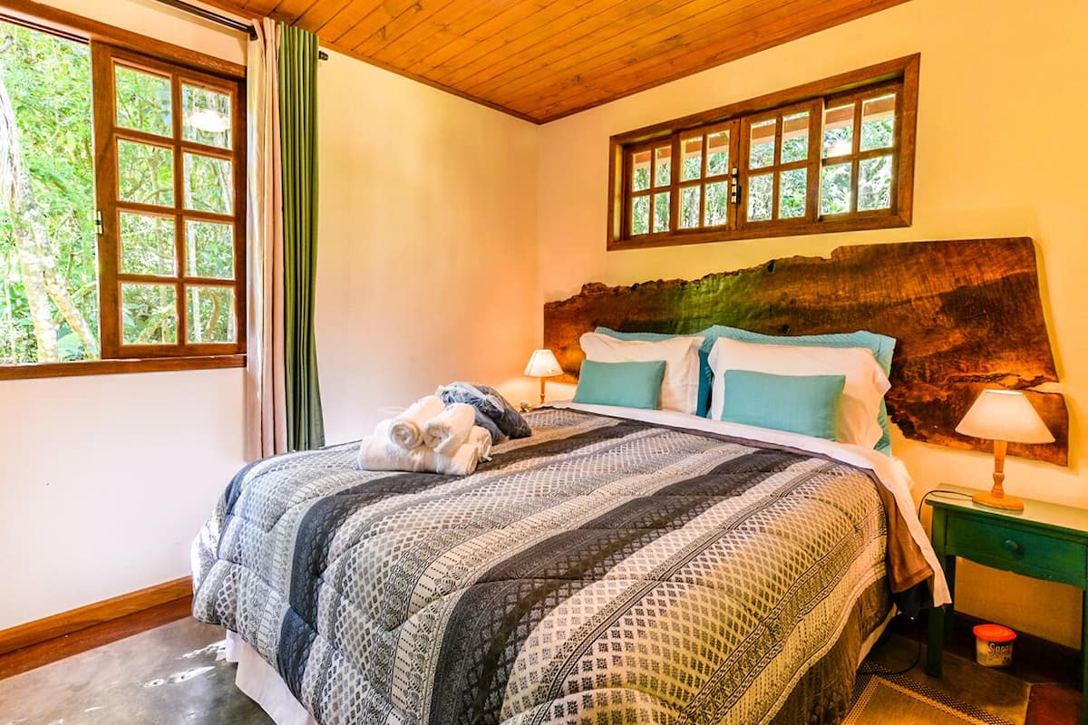 Une chambre à coucher avec un grand lit, une tête de lit en bois, deux tables de chevet avec des lampes et une vue sur la verdure par la fenêtre.