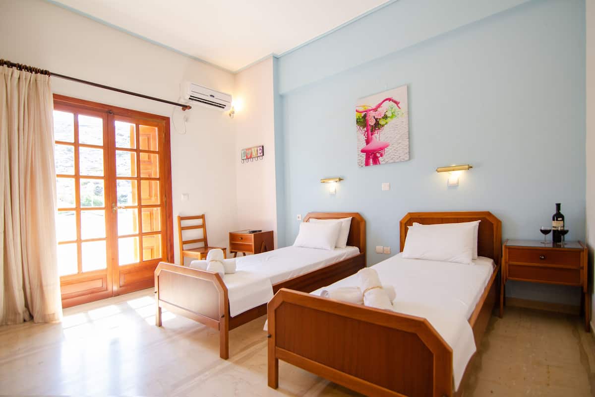 Une chambre d’hôtel avec deux lits en bois, un bureau, une chaise, une fenêtre avec des rideaux, un climatiseur et une œuvre d’art accrochée au mur.