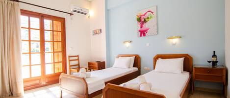 A hotel room with two wooden beds, a desk, a chair, a window with curtains, an air conditioning unit, and a wall-mounted artwork.