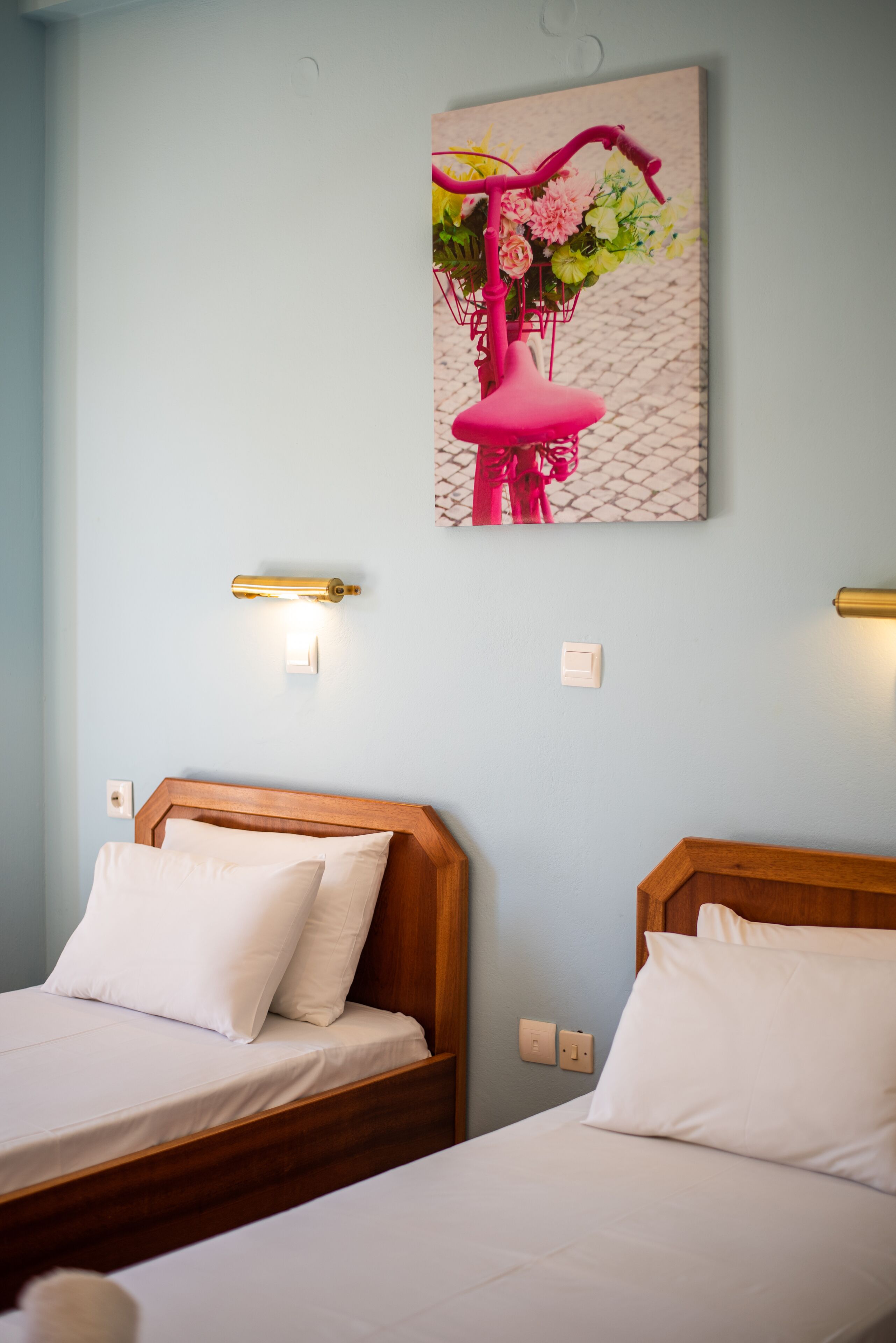 A hotel room with two beds, light blue walls, and a painting of a pink bicycle with flowers.