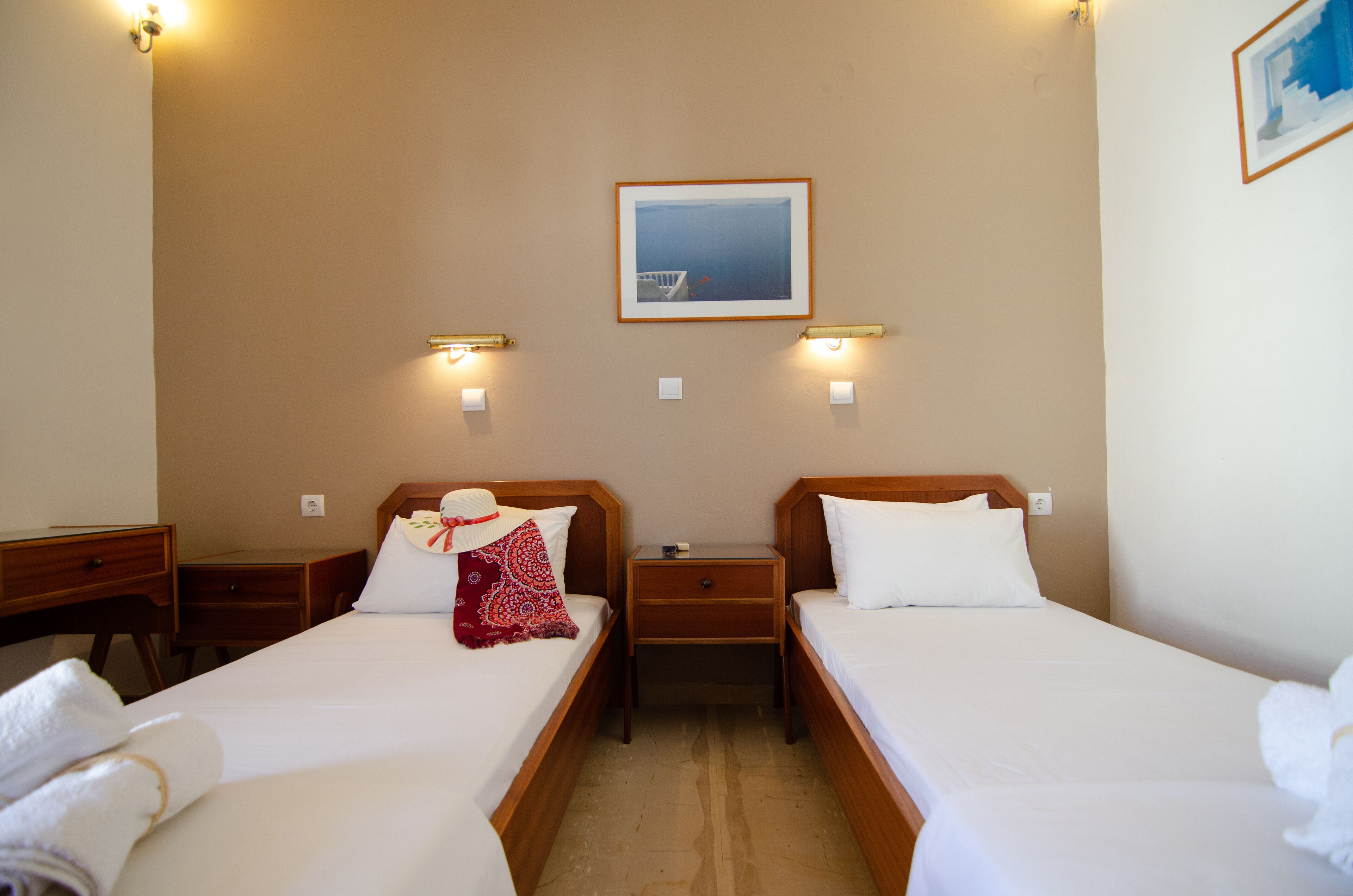A hotel room with two single beds, a desk, and a nightstand. There is a framed picture on the wall and wall-mounted lights.