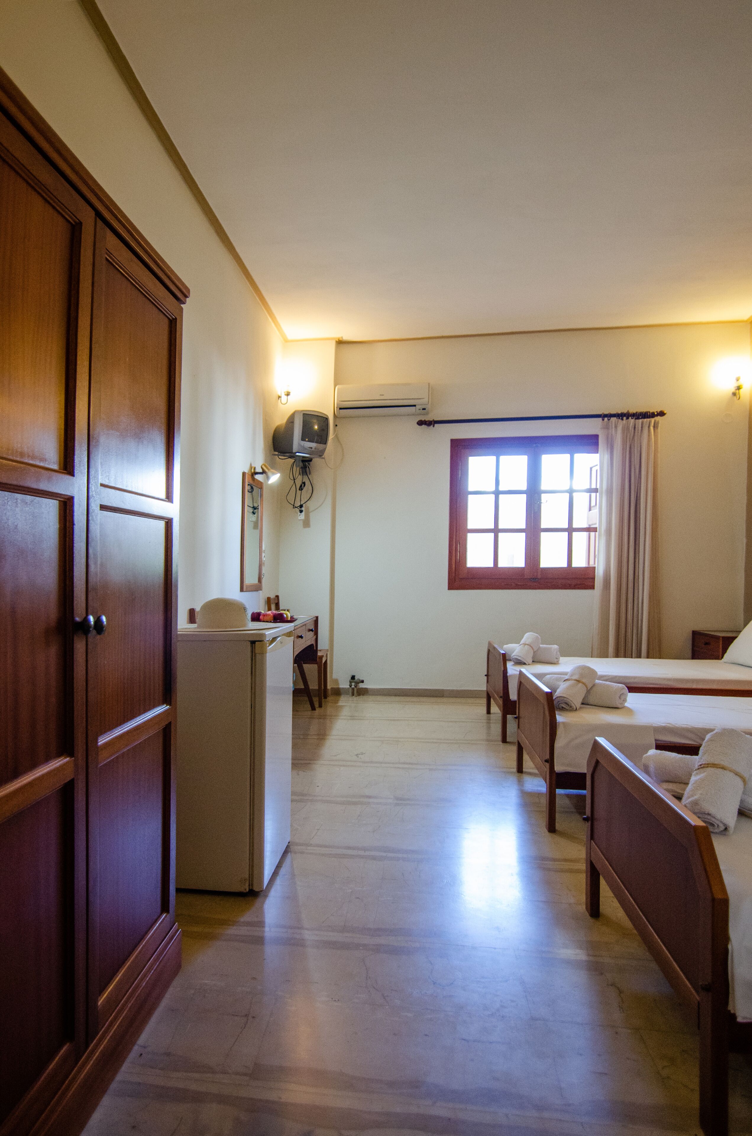 Una camera d'albergo con un armadio in legno, una scrivania con una sedia, un letto con due cuscini, una finestra con tende e un televisore fissato al muro.