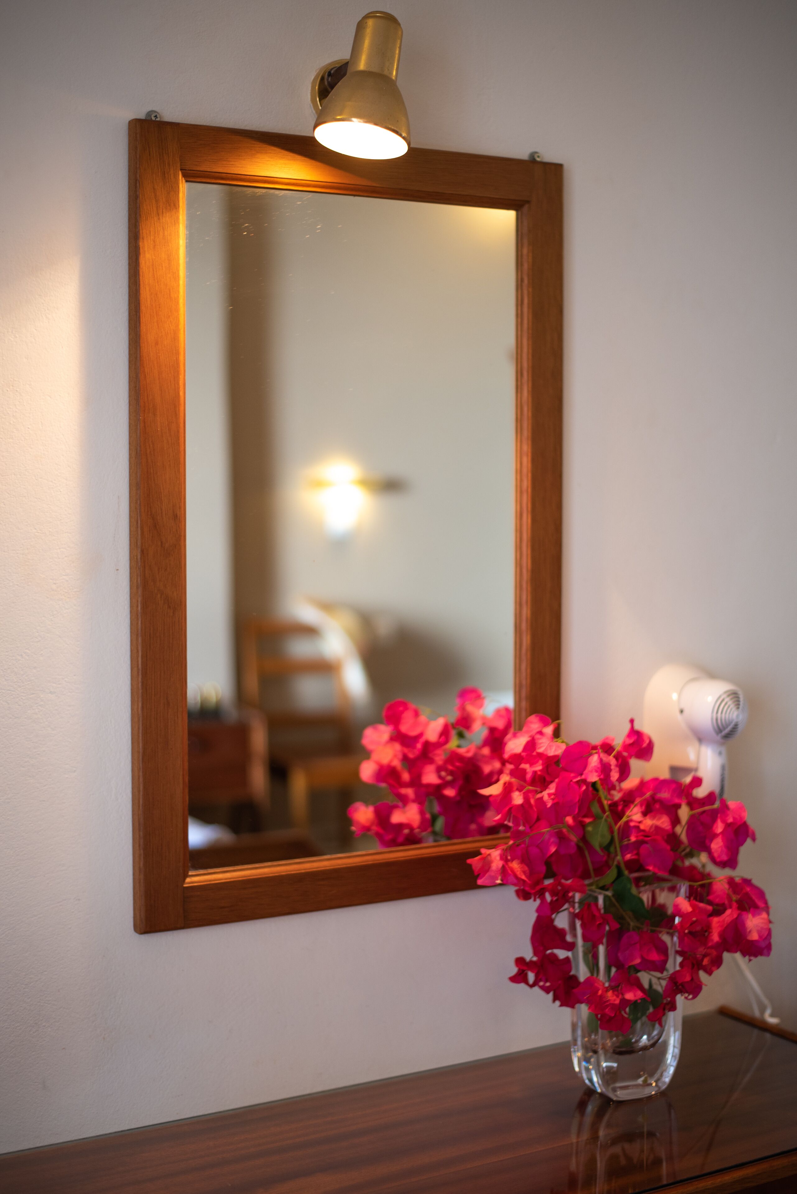 A mirror with a wooden frame, a hairdryer on a wooden surface, and a vase with pink flowers.