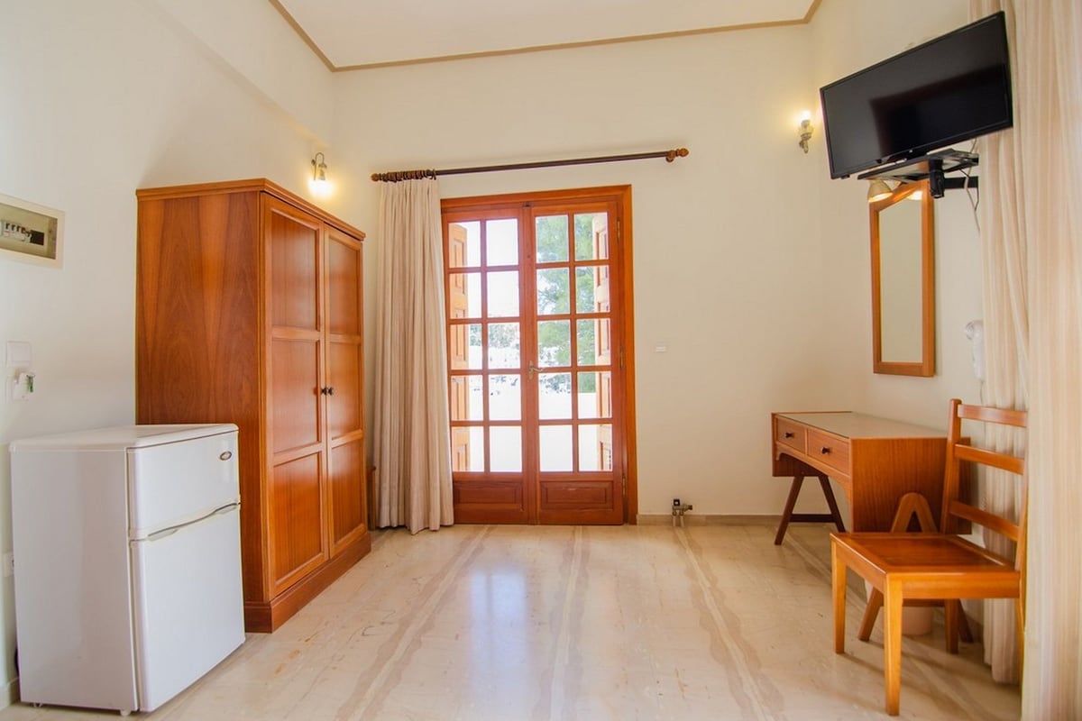 A room with a wooden wardrobe, a desk with a chair, a television mounted on the wall, and a window with curtains.