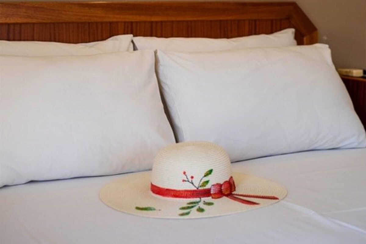 A hotel room with a bed, white pillows, a white hat on the bed, and a wall-mounted light fixture.