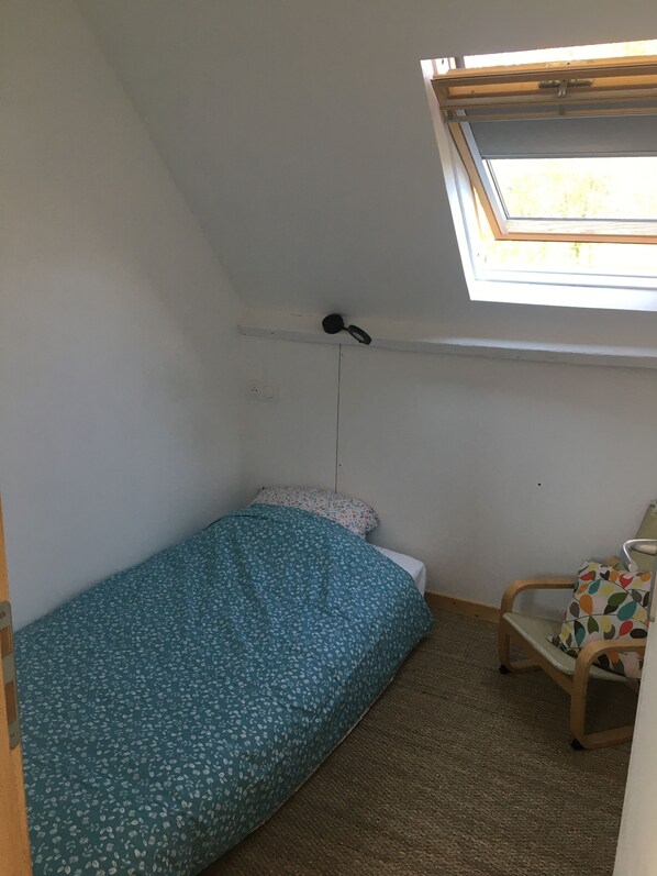 A small, single-bed room with a skylight, a chair, and a power outlet on the wall.
