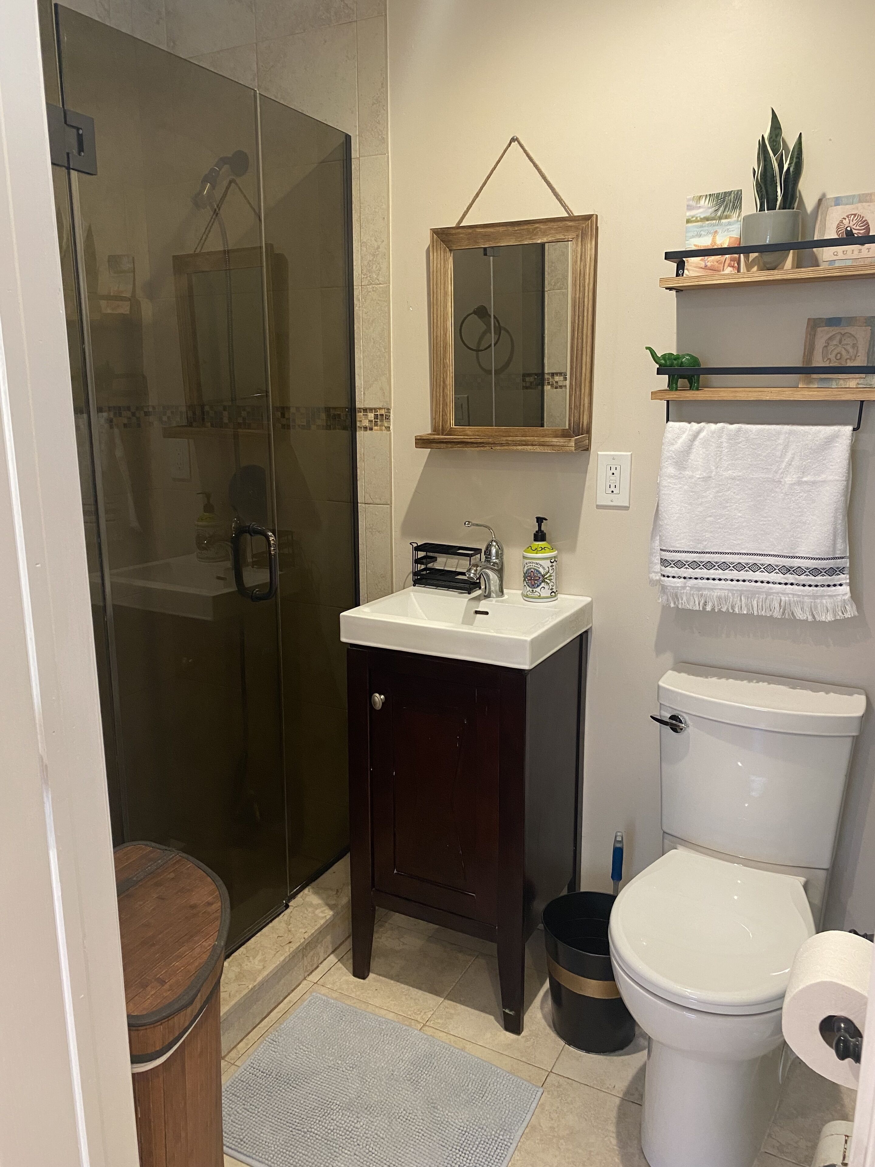 Une salle de bain avec une douche à parois de verre, un lavabo blanc avec un meuble en bois, des toilettes et une étagère ornée d’objets décoratifs.