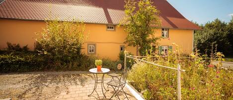 A yellow house with a red roof, a paved patio with a table and chairs, and a garden with various plants and flowers.