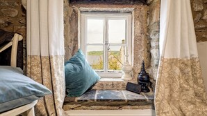 Una habitación rústica con pared de piedra, techo de madera, ventana con vistas, un escalón pequeño que lleva a la ventana, una lámpara blanca y un cojín azul.