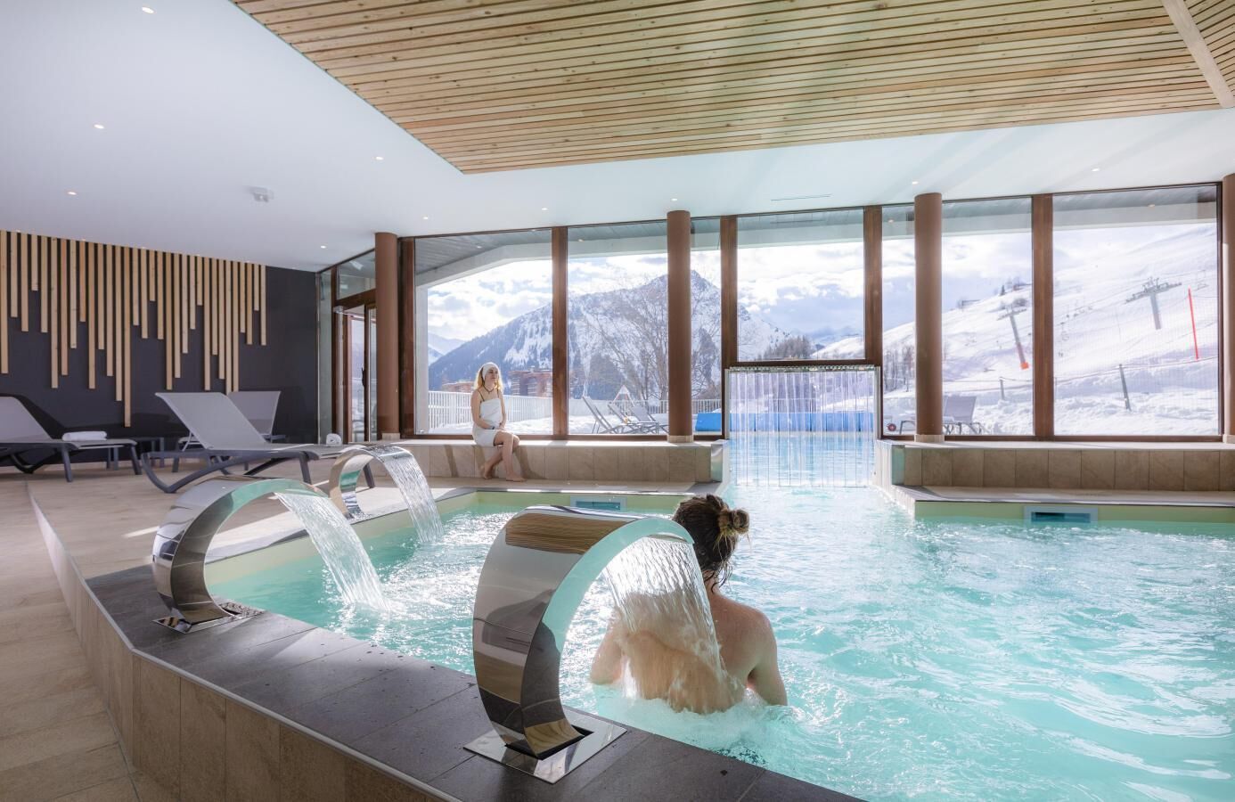 A modern indoor pool with a view of snowy mountains, lounge chairs, and a wooden ceiling.
