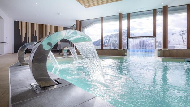 Une piscine intérieure moderne dotée d’un élément aquatique incurvé original et de grandes fenêtres offrant une vue sur les montagnes enneigées.