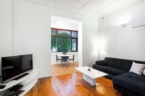 A modern living room with a flat-screen TV, a dark grey sofa, a white coffee table, and a dining area with a table and chairs.