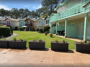 A courtyard with a green lawn, potted plants, and a building with a balcony. - Nice house in residence with large swimming pool (Lacanau)