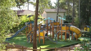 A playground with slides and climbing structures surrounded by trees and a wooden fence.
