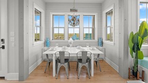 Un coin repas avec une table blanche dressée pour quatre personnes, entourée de chaises grises, de grandes fenêtres et d’un lustre.
