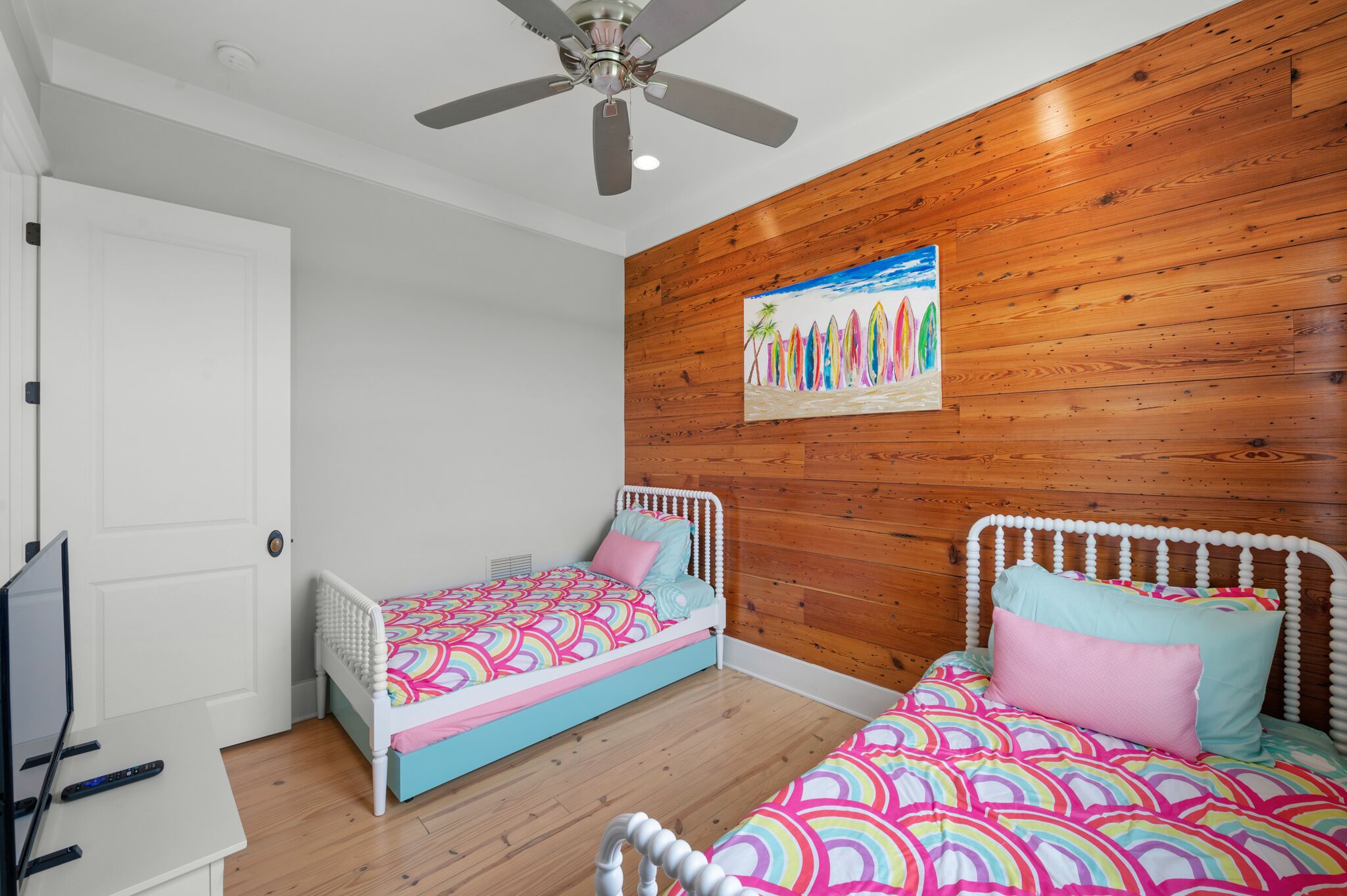 A bedroom with two beds, wooden paneling on the walls, a ceiling fan, and a painting of surfboards.