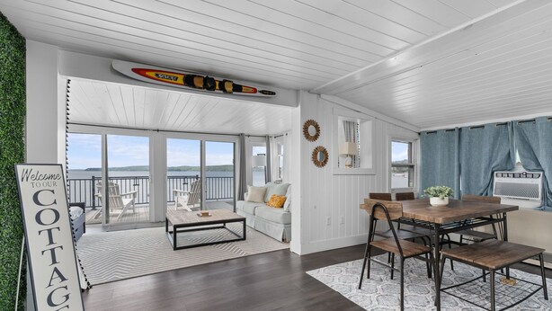 A spacious living area with a large window, a wooden dining table, and a couch. - Lakeside Cottage ~ Pool, Hot tub & Private Beach (Colchester)
