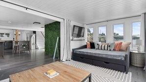 A modern living room with a sofa, a coffee table, and a TV. There is a staircase leading to a kitchen area. - Lakeside Cottage ~ Pool, Hot tub & Private Beach (Colchester)