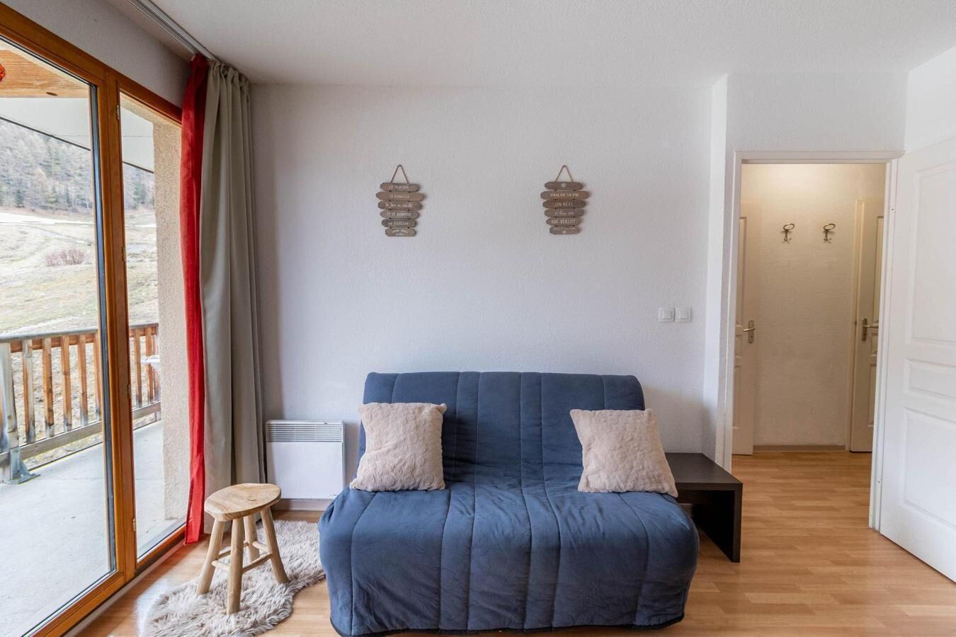 Una habitación con un sofá azul, dos almohadas, un taburete pequeño, un radiador y vistas a un paisaje nevado a través de un ventanal grande.
