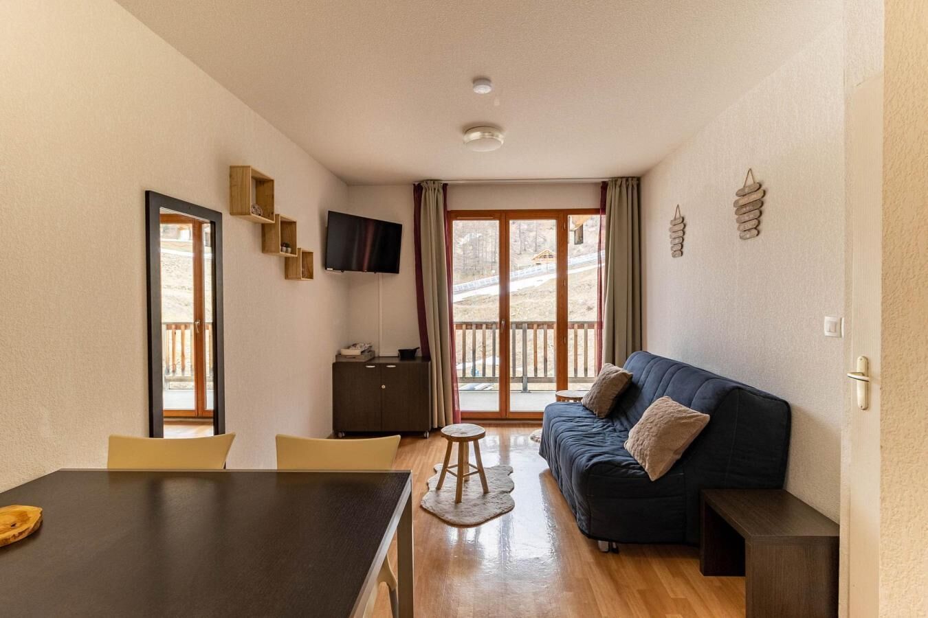 Un espacio habitable compacto con un sofá oscuro, una pequeña mesa de comedor, un televisor montado en la pared y un ventanal corredero que da a un área exterior.