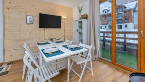A dining area with a white table and chairs, a flat-screen TV mounted on the wall, and a sliding glass door leading to a balcony with a view of buildings. - 200m from ski slopes, 1st floor, view resort, swimming pool, balcony, tv, ski locker, 24m² (Les Orres)