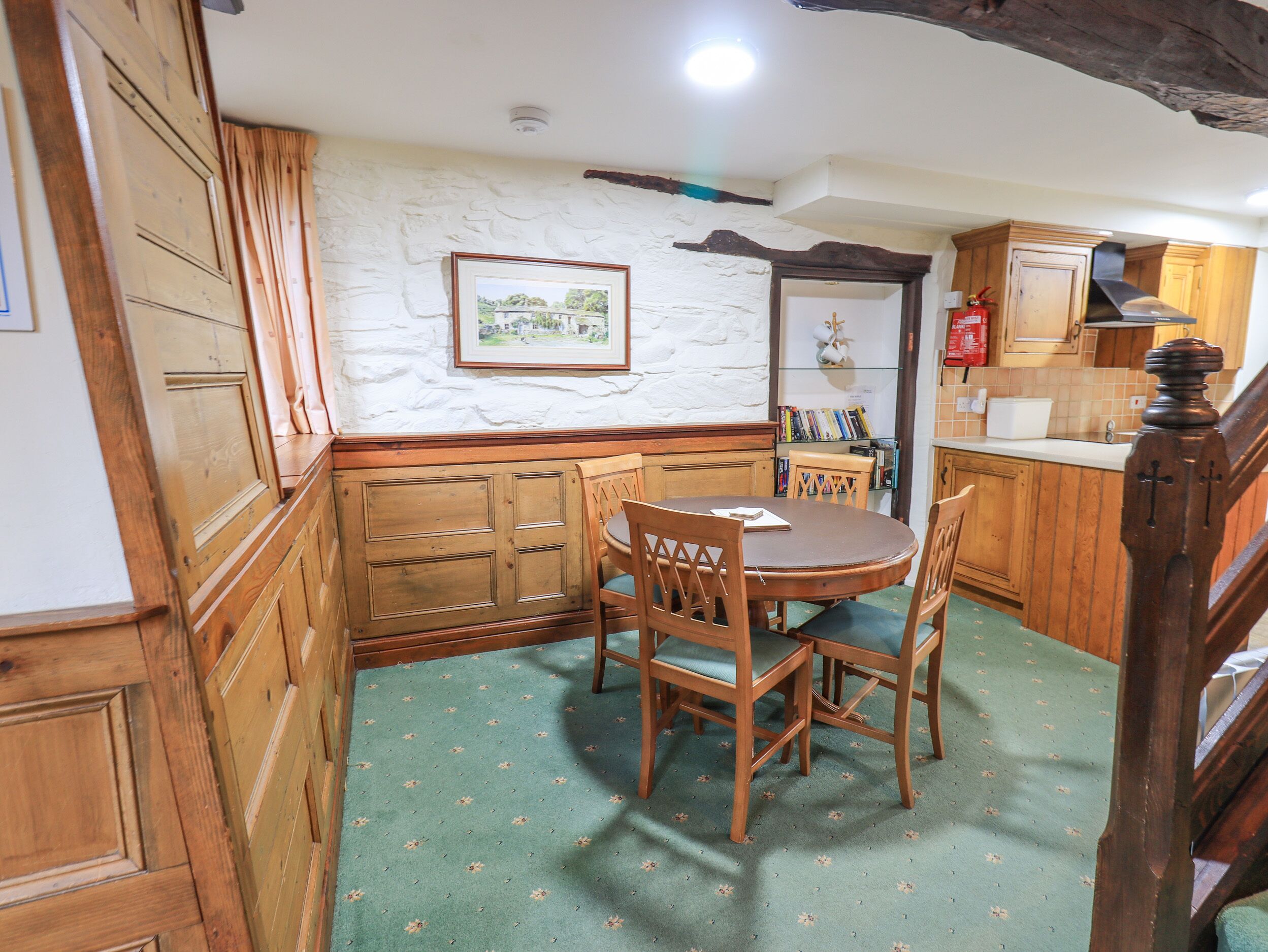 Un área de comedor con una mesa redonda de madera y sillas, una alfombra verde estampada, armarios de madera y un cuadro en la pared.