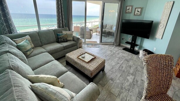 A living room with a large window overlooking the beach, a flat-screen TV, and a wicker chair. - Silver Beach 504 | Updated, Direct Beach Front, Pet Friendly! (Orange Beach)