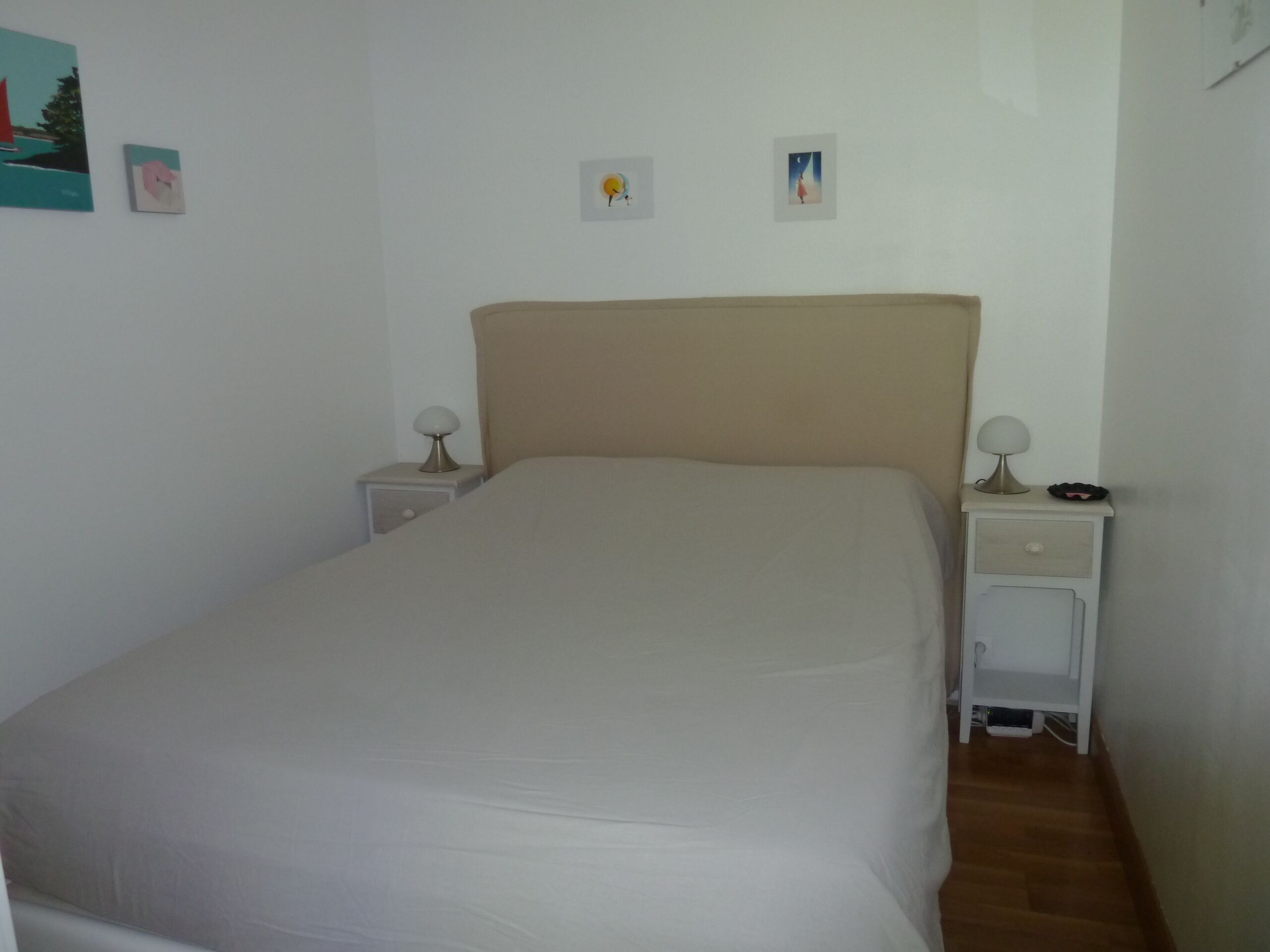 A neatly made bed in a room with a plain white wall, two bedside tables with lamps, and a small shelf under the right table.