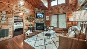 Interior de una cabaña de troncos con chimenea, televisor montado en una pared de piedra, una cama, un sofá y una mesa de comedor.