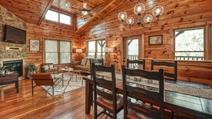 Interior acogedor de una cabaña con paredes de madera, una chimenea de piedra, una zona de comedor con sillas y una mesa, y un ventilador de techo.