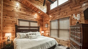 Un dormitorio con paneles de madera, una cama, mesitas de noche, un aparador y un ventilador de techo.
