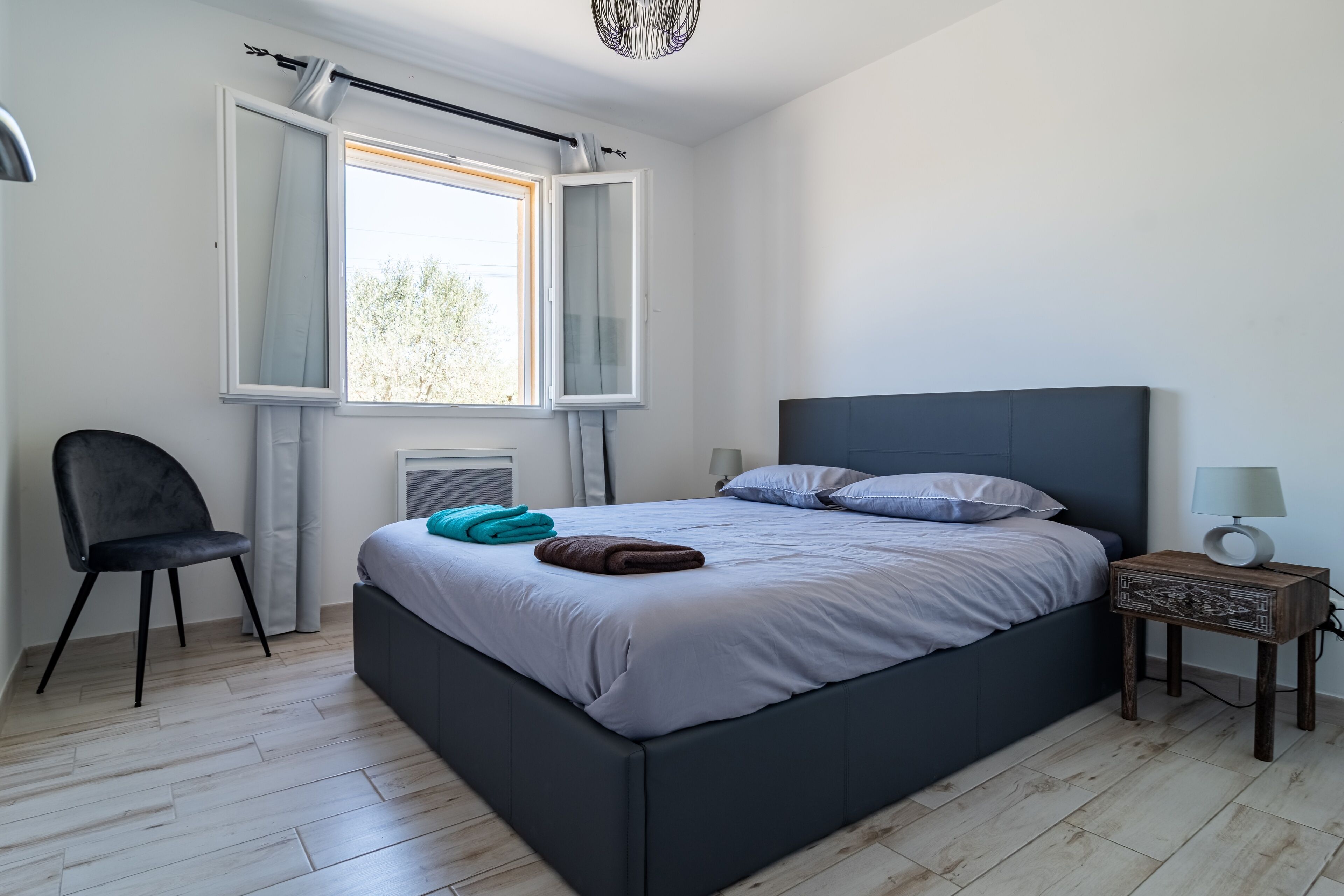 A modern bedroom with a large bed, a chair, a nightstand, and a window with curtains.
