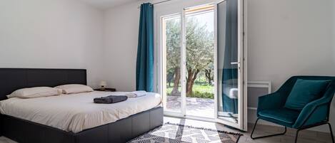 A bedroom with a bed, a chair, a patterned rug, and a view through a glass door.