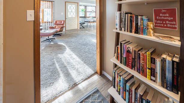 Un couloir avec une bibliothèque remplie de livres, un ventilateur de plafond et un tapis menant à une pièce où se trouvent un bureau et des chaises.