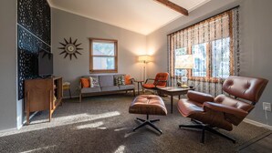 Un salon comprenant un canapé, un fauteuil, un pouf et un téléviseur posé sur un meuble en bois.
