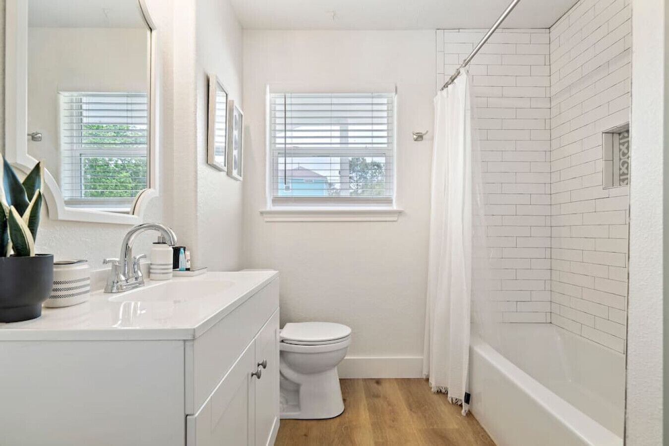 A bathroom with a white sink, mirror, and toilet. There are two windows with blinds, and a shower area with white tiles.