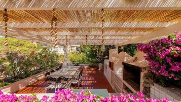 Relax in a shaded patio surrounded by lush greenery and vibrant flowers, complete with a grill and comfortable seating.