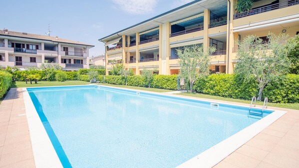 Une piscine rectangulaire bordée de blanc, entourée d’une terrasse carrelée. On aperçoit, en arrière-plan, des immeubles à plusieurs étages dotés de balcons et un arbuste taillé avec soin.