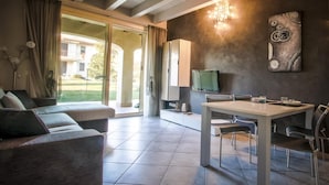 A modern living room with a sofa, a dining table with chairs, a TV, and a large window with curtains. - La Moderna Egman - Italian Homing (Sirmione)