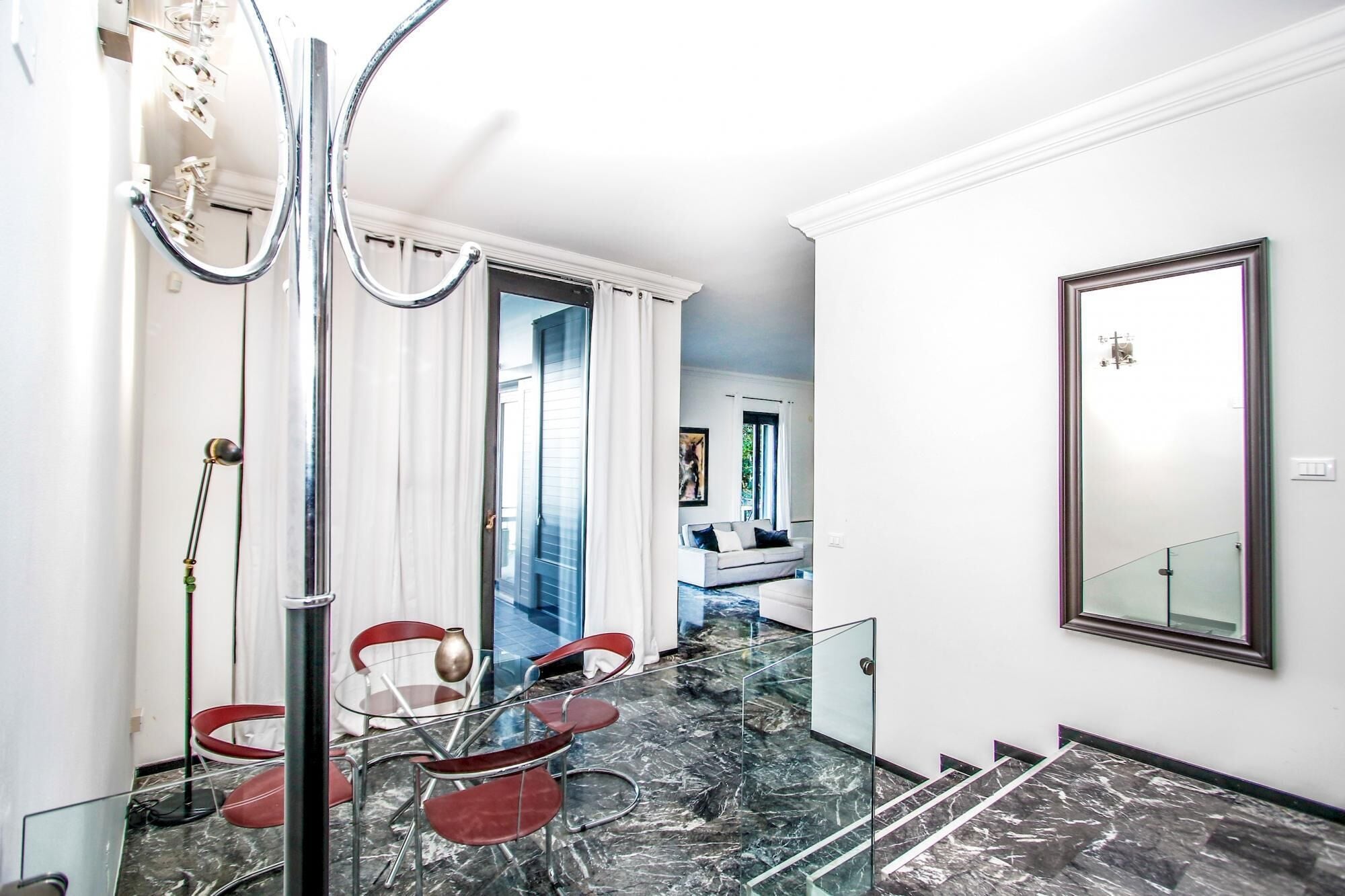 Interior moderno com escada de vidro, área de jantar com cadeiras vermelhas e um espelho grande na parede.