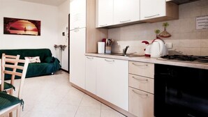Cocina compacta con armarios blancos, un fregadero, estufa y lavavajillas. En la zona de estar contigua, hay un sofá verde y una silla de madera.