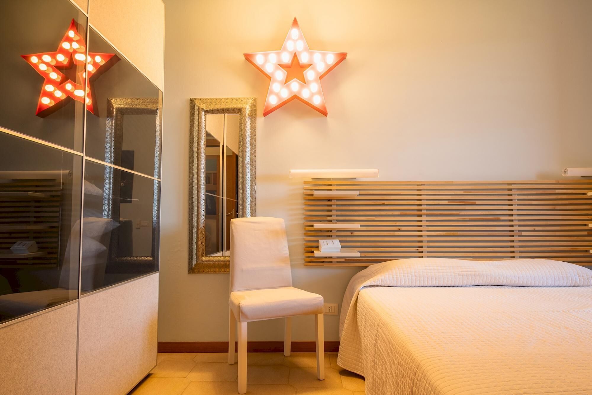 A hotel room with a bed, a chair, a mirror, and two illuminated star decorations on the wall.